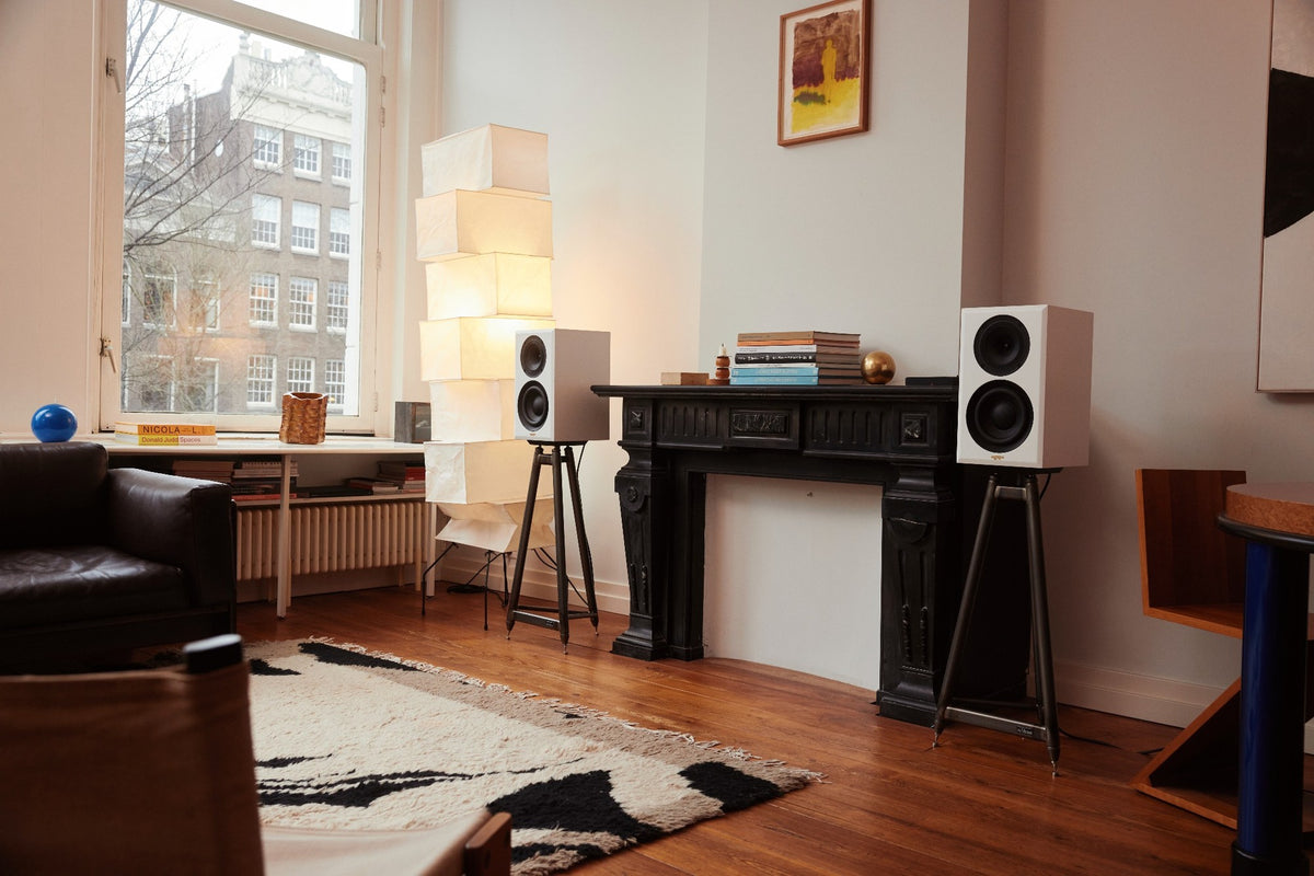 A pair of white True Tone speakers on tripod stands beside a fireplace, with a paper lantern and a window behind